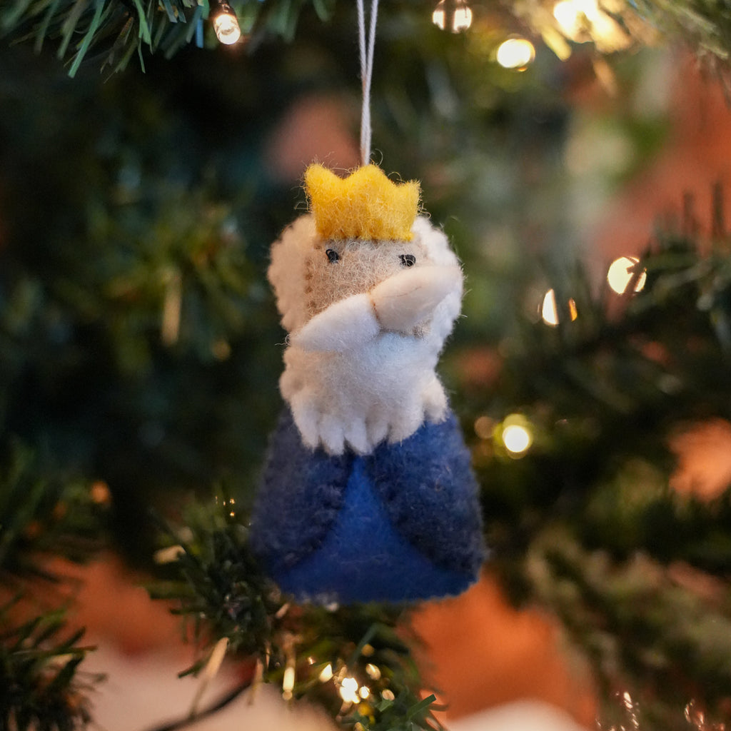 A small felt Christmas ornament shaped like a crowned figure with a white beard and blue lower body, hanging on a Christmas tree branch with warm lights in the background.