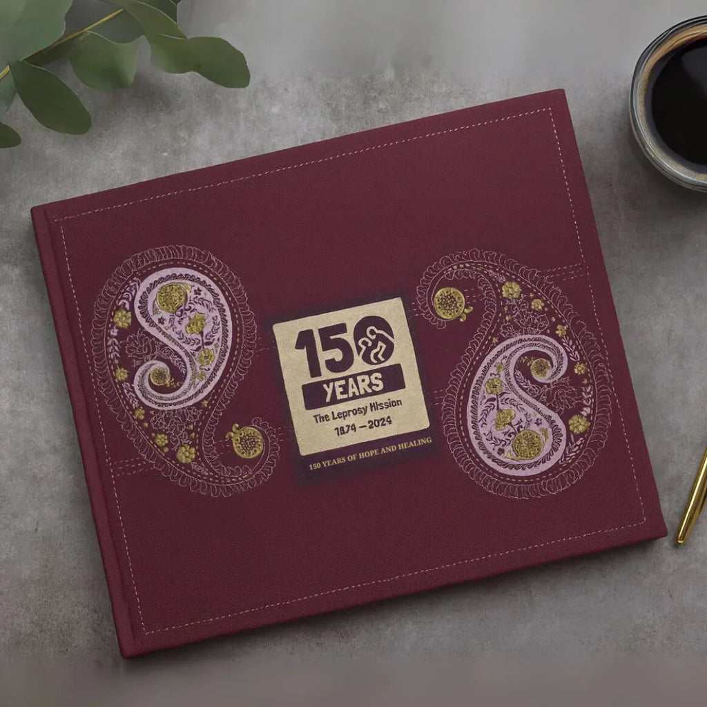 Purple book with paisley design and '150 Years' label on a gray surface with a plant and coffee cup.