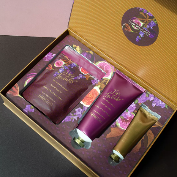 A luxury gift set of three bath and body products from The Lavender Company, presented in an open gold box with floral purple lining. The set includes a hand wash, a body lotion tube, and a hand cream tube, all decorated with purple lavender and pink rose illustrations.