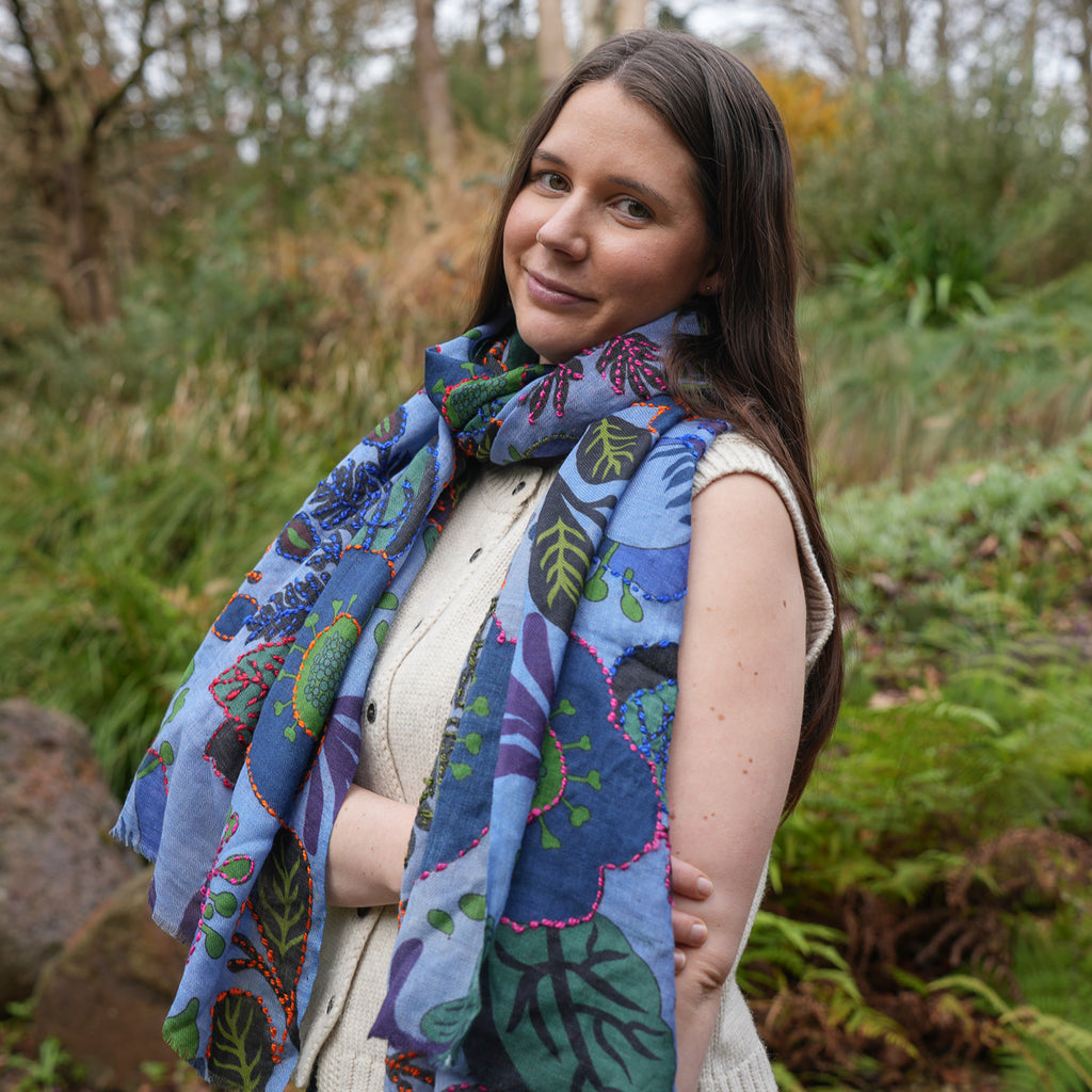 A woman wearing a vibrant blue patterned scarf with botanical and abstract designs in green, purple, and pink, set against a blurred outdoor background of green foliage.