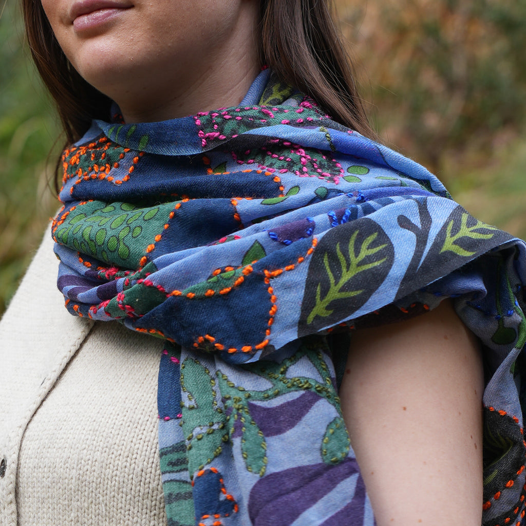 A close-up of a woman wearing a blue patterned scarf with green, purple, and orange botanical designs, paired with a beige knit cardigan.