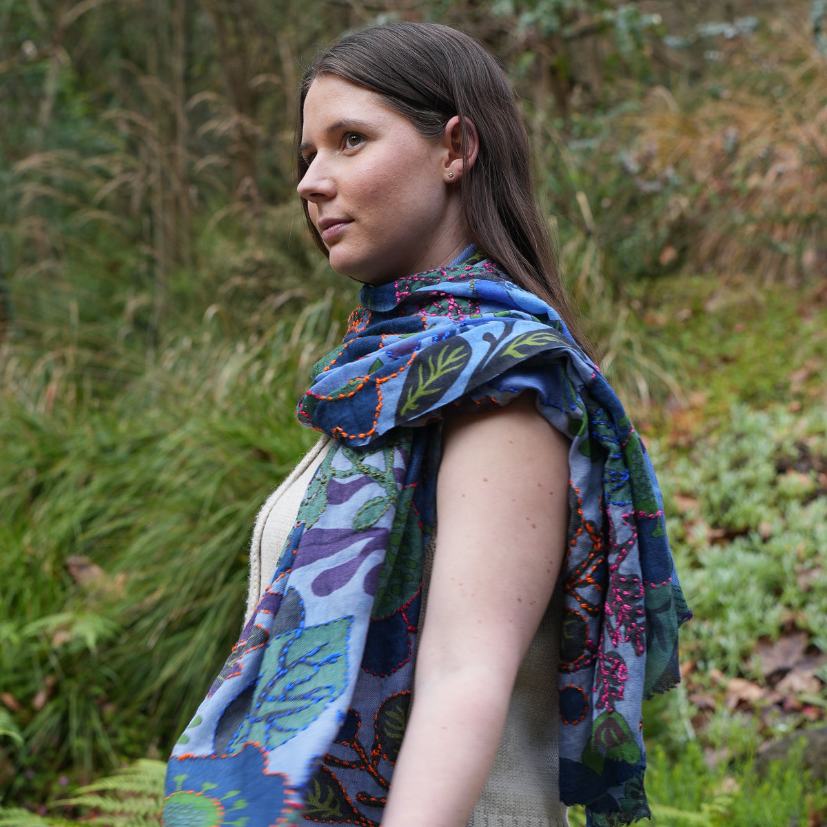 A woman wearing a blue patterned scarf with green, purple, and red botanical designs, standing outdoors in a grassy, green environment.