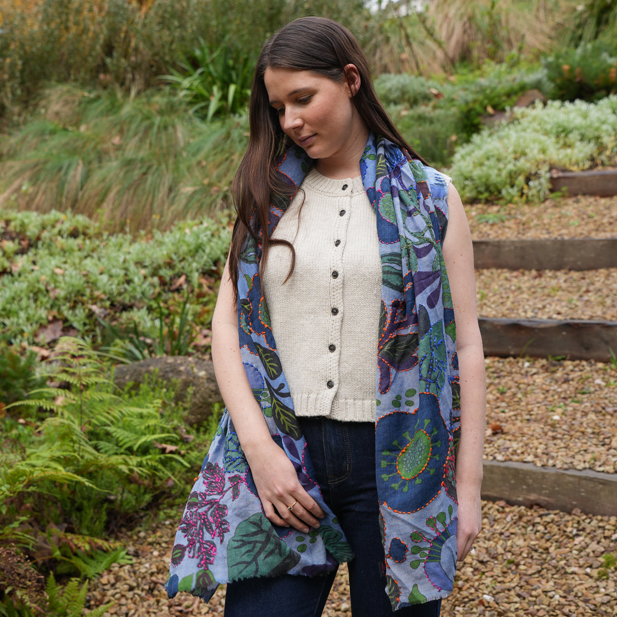 A woman standing outdoors in a garden wearing a sleeveless cream knit cardigan vest over dark jeans, with a blue and green paisley patterned scarf draped around her neck.