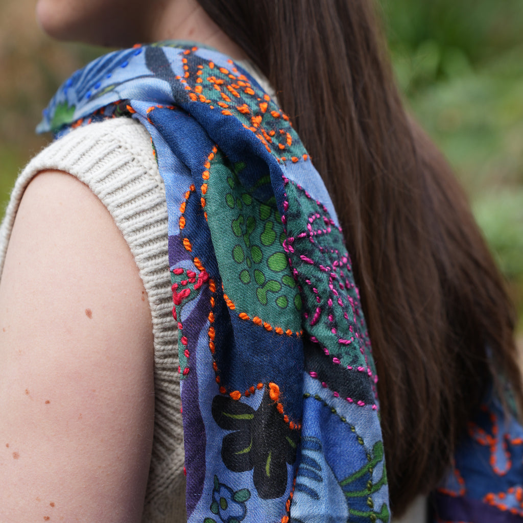 A close-up lifestyle shot of a woman wearing a blue patterned scarf with green, orange, and pink embroidered details over a beige ribbed sleeveless top, with a blurred green outdoor background.