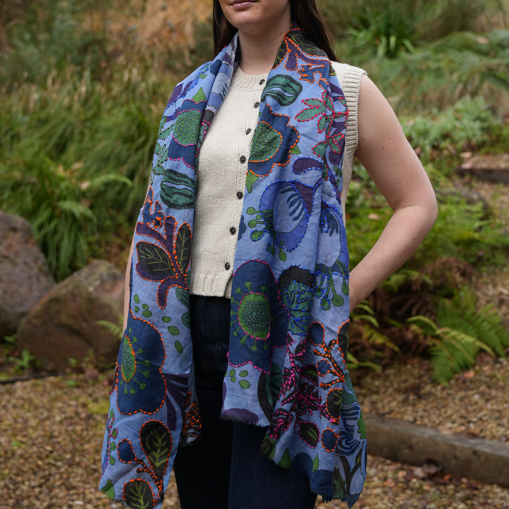 A woman wearing a light blue patterned scarf with green, purple, and red botanical motifs, styled over a cream sleeveless top in an outdoor garden setting.