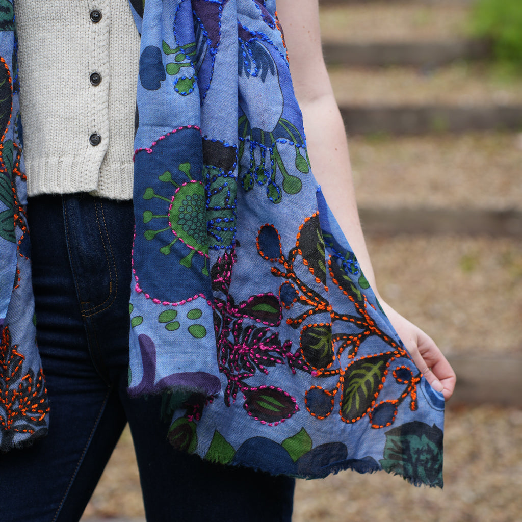 A person outdoors wearing a light beige knit cardigan and dark blue jeans, holding up a blue patterned scarf with botanical and coral-like motifs in green, orange, purple, and pink.