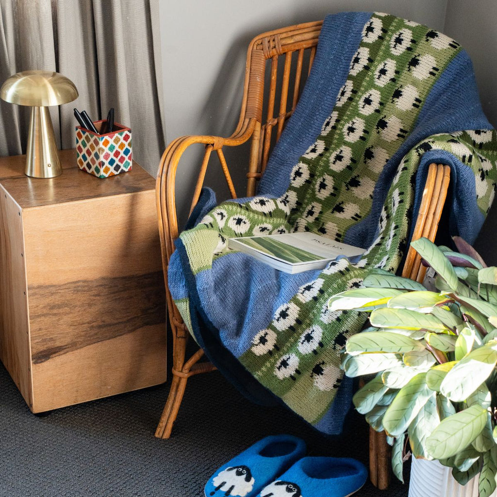 Wicker chair with a floral-patterned blanket draped over it, next to a wooden side table and plant.