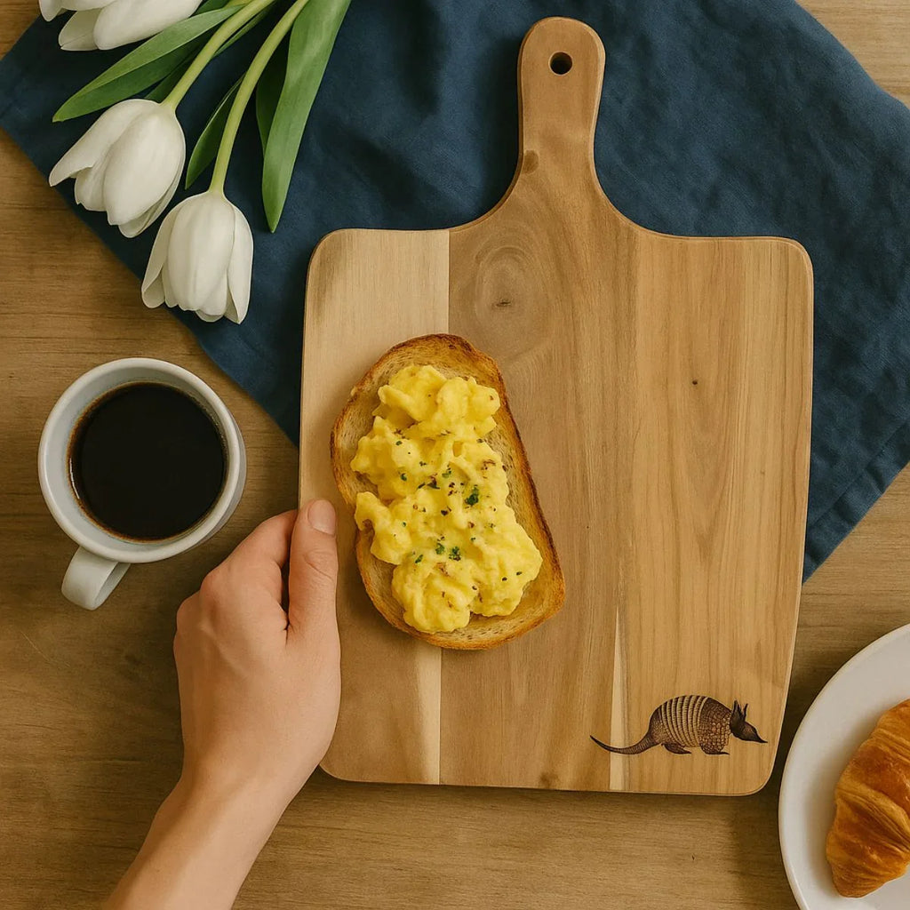 Wooden cutting board with scrambled eggs on toast, a cup of coffee, and tulips on a wooden table.