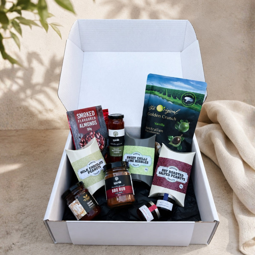 An open white gift box filled with assorted gourmet snacks and treats, photographed outdoors on a beige surface with soft shadows and a draped fabric beside it. Visible items include packaged almonds, a jar of bad rub, cheese wedges, and other snack products.