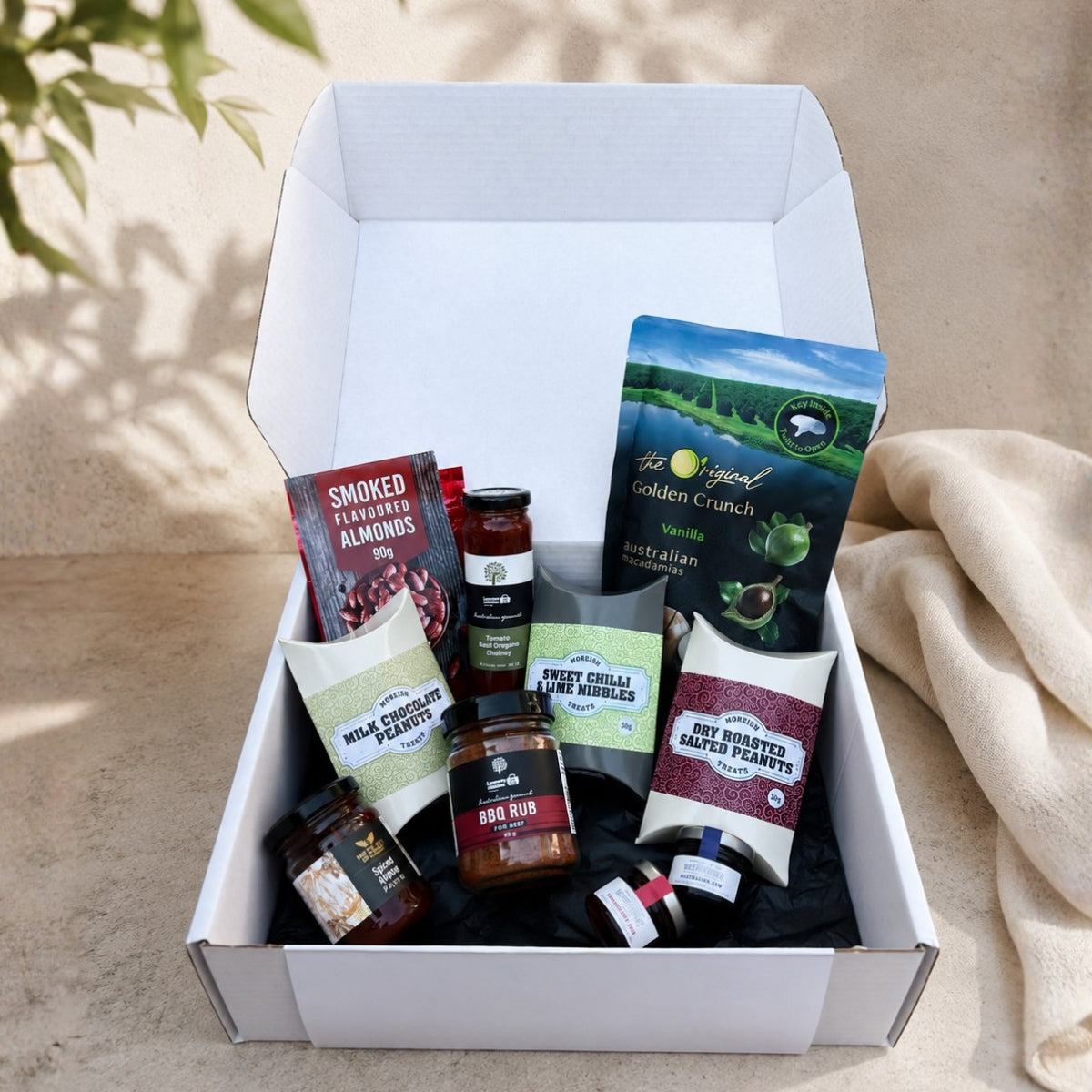 An open white gift box filled with assorted gourmet snacks and treats, photographed outdoors on a beige surface with soft shadows and a draped fabric beside it. Visible items include packaged almonds, a jar of bad rub, cheese wedges, and other snack products.
