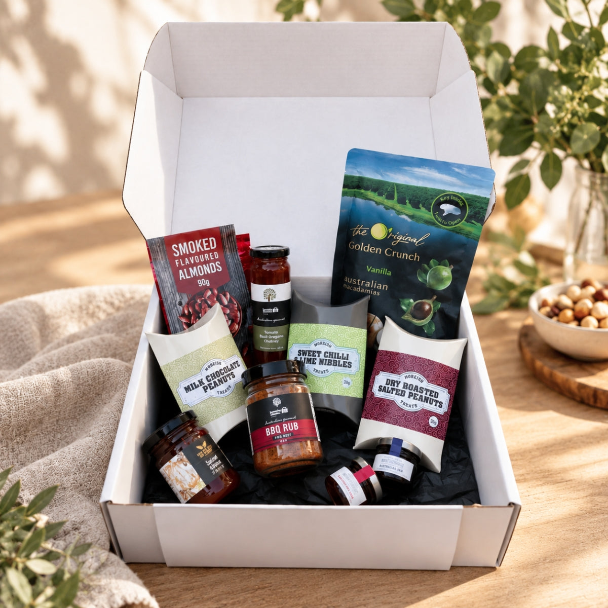 An open white gift box filled with assorted gourmet snacks and treats, photographed on a wooden table with natural sunlight and blurred greenery in the background. Visible items include bags of chips, jars of spread, nuts, and packaged snacks.