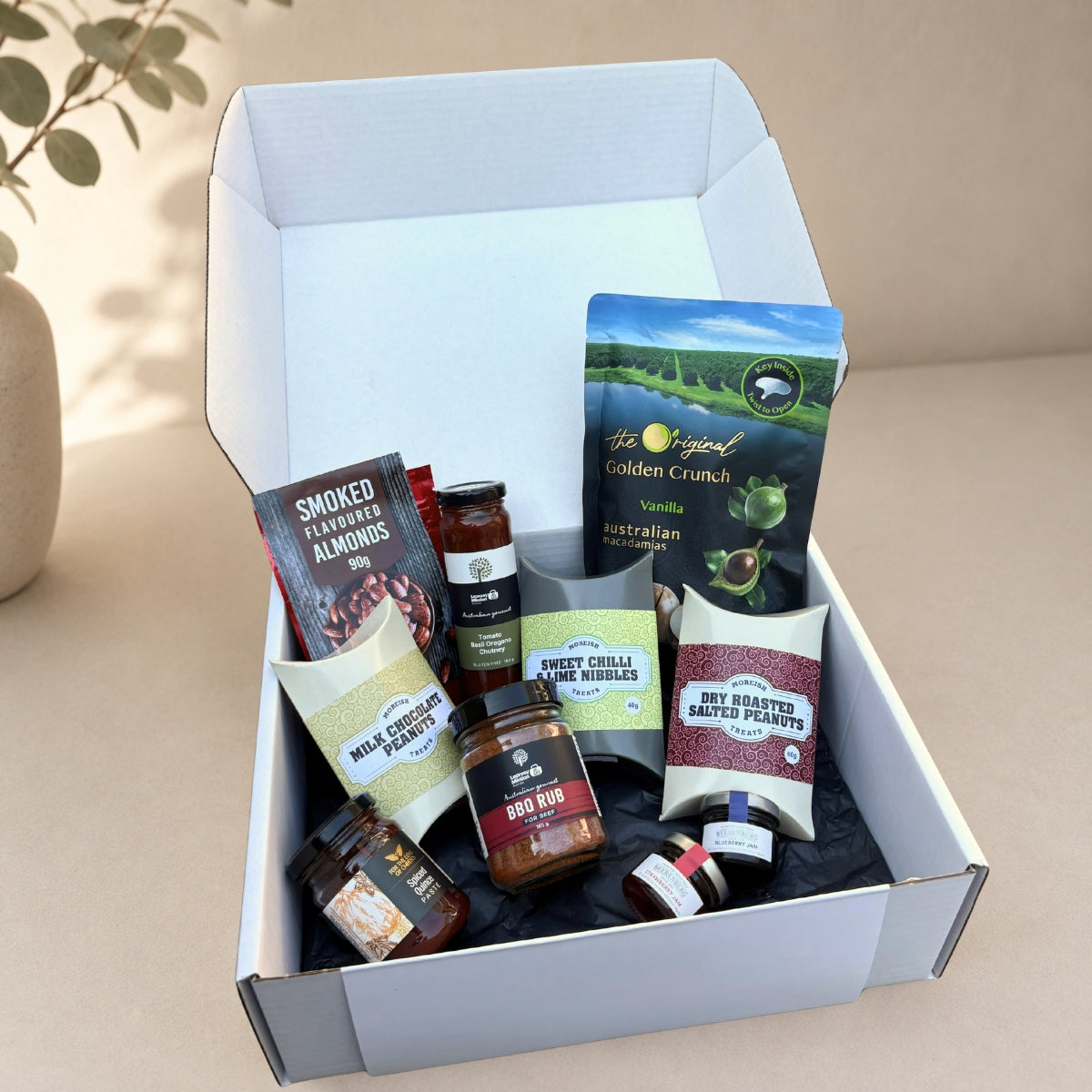 An open white gift box filled with assorted packaged food items, including a bag of Golden Crunch chips, a jar of BBQ rub, a bag of roasted garlic peanuts, and other snack packages, photographed on a beige surface with a soft neutral background.