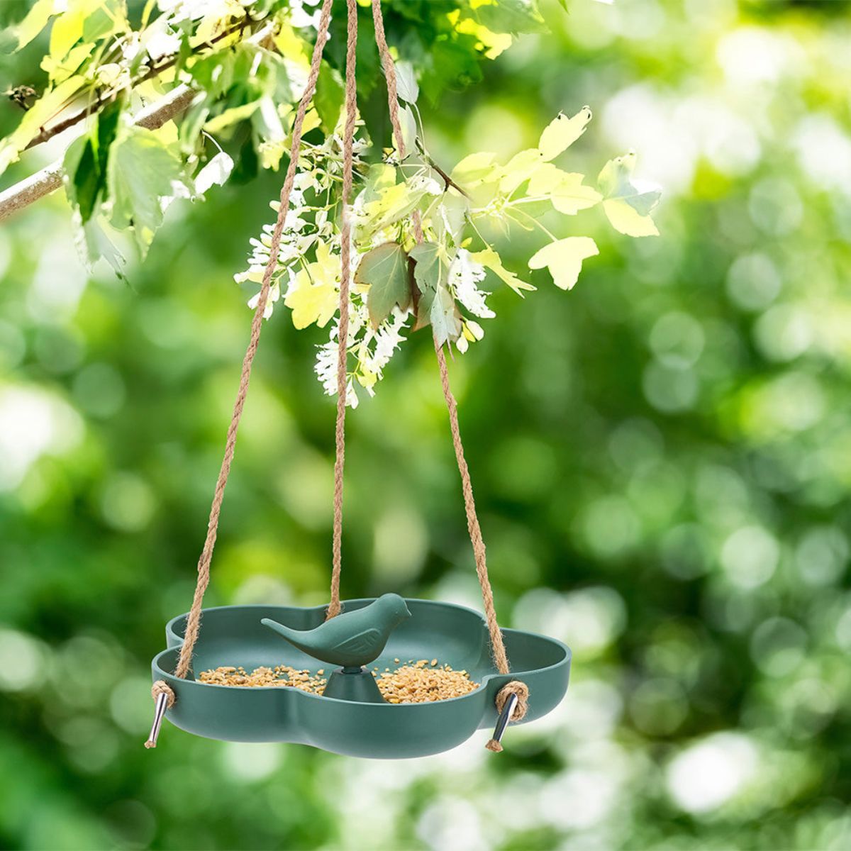 Green bird feeder hanging from a branch with a blurred green foliage background