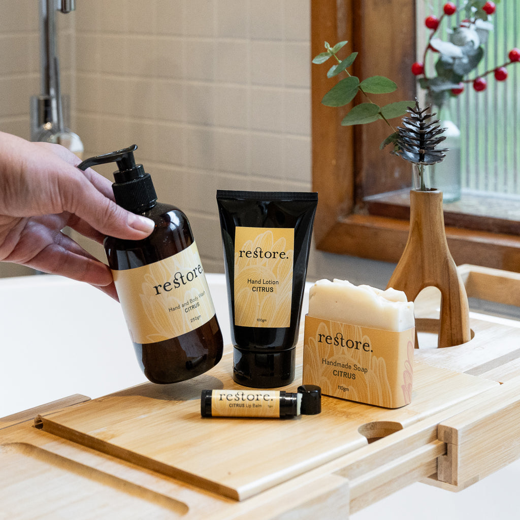 A hand holds a dark amber bottle of Restore Hand & Foot Cream next to a black tube and a bar of handmade soap on a wooden bath tray in a bathroom setting.