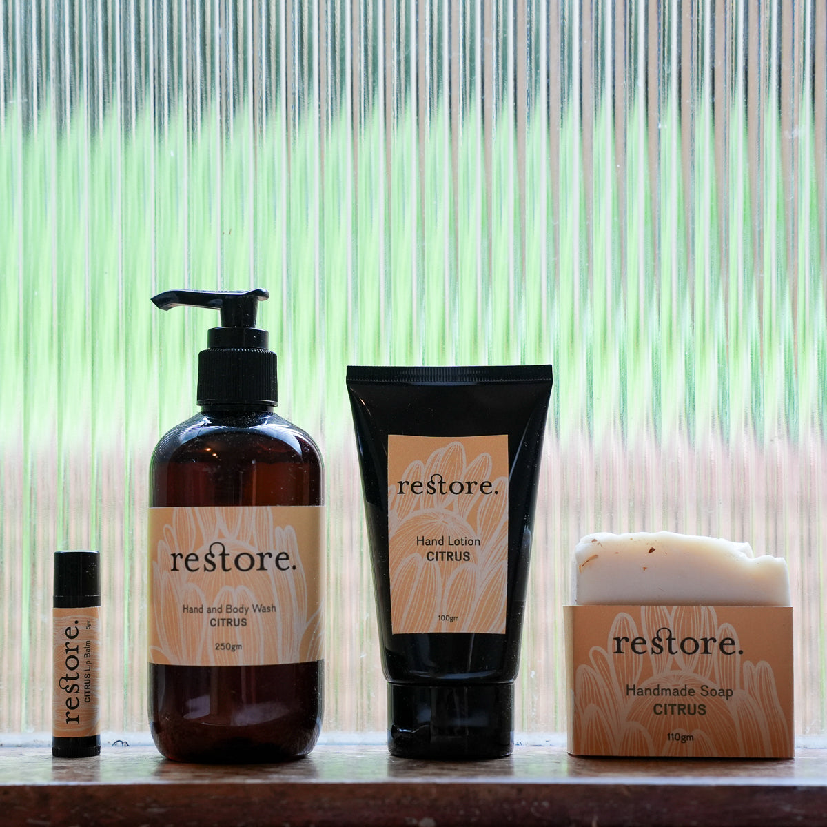 Four Restore brand self-care products arranged on a shelf in front of a green and white striped curtain: a hand and body wash pump bottle, a hand lotion squeeze tube, a small body butter or perfume roller bottle, and a jar of citrus handcrafted soap.