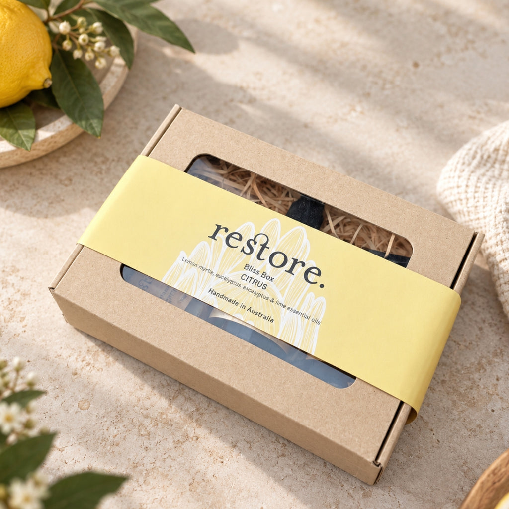 A kraft paper gift box with a yellow paper band reading 'restore. Bliss Box CITRUS' and 'Handmade in Australia,' photographed on a light stone surface with a lemon and green leaves nearby.
