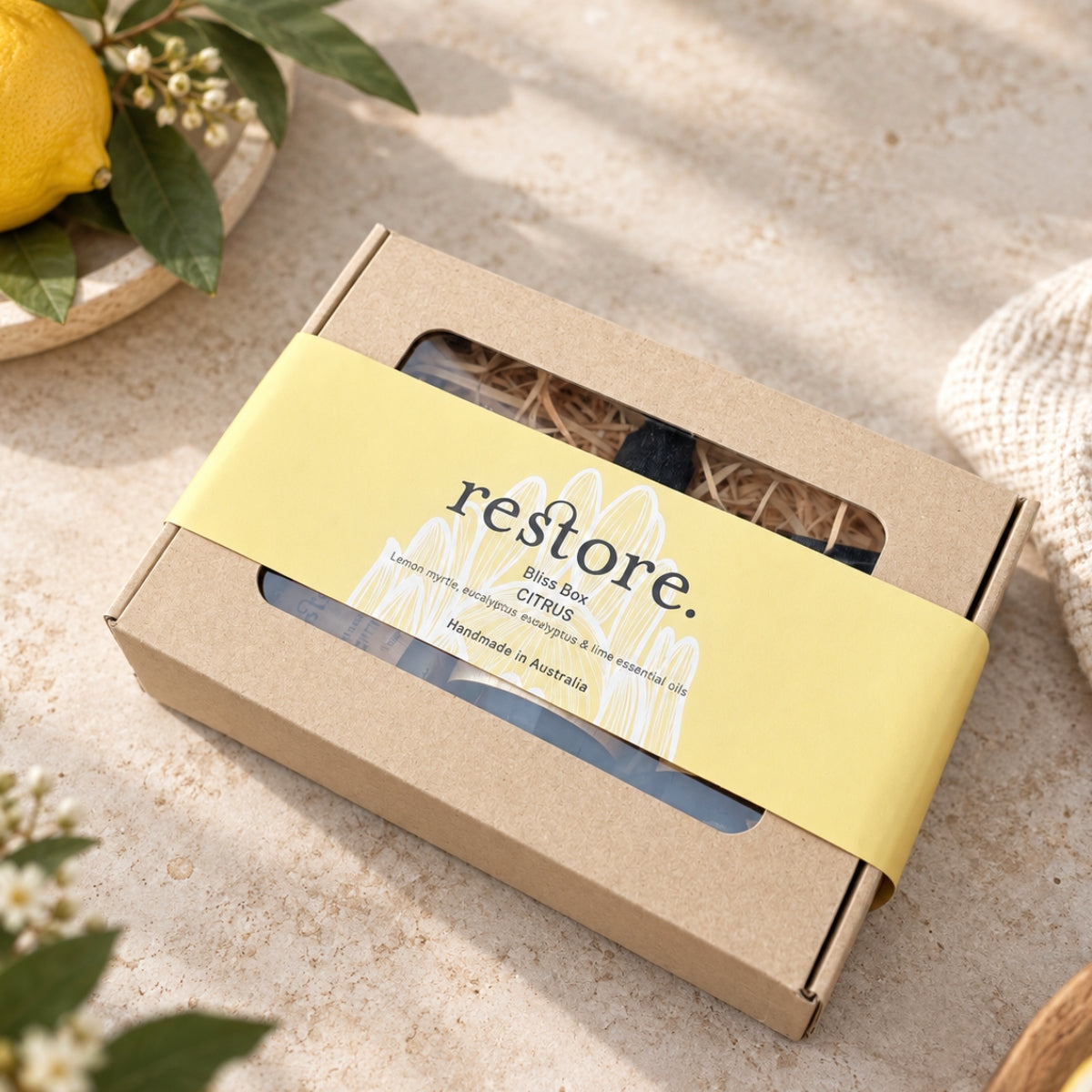 A kraft paper gift box with a yellow paper band reading 'restore. Bliss Box CITRUS' and 'Handmade in Australia,' photographed on a light stone surface with a lemon and green leaves nearby.