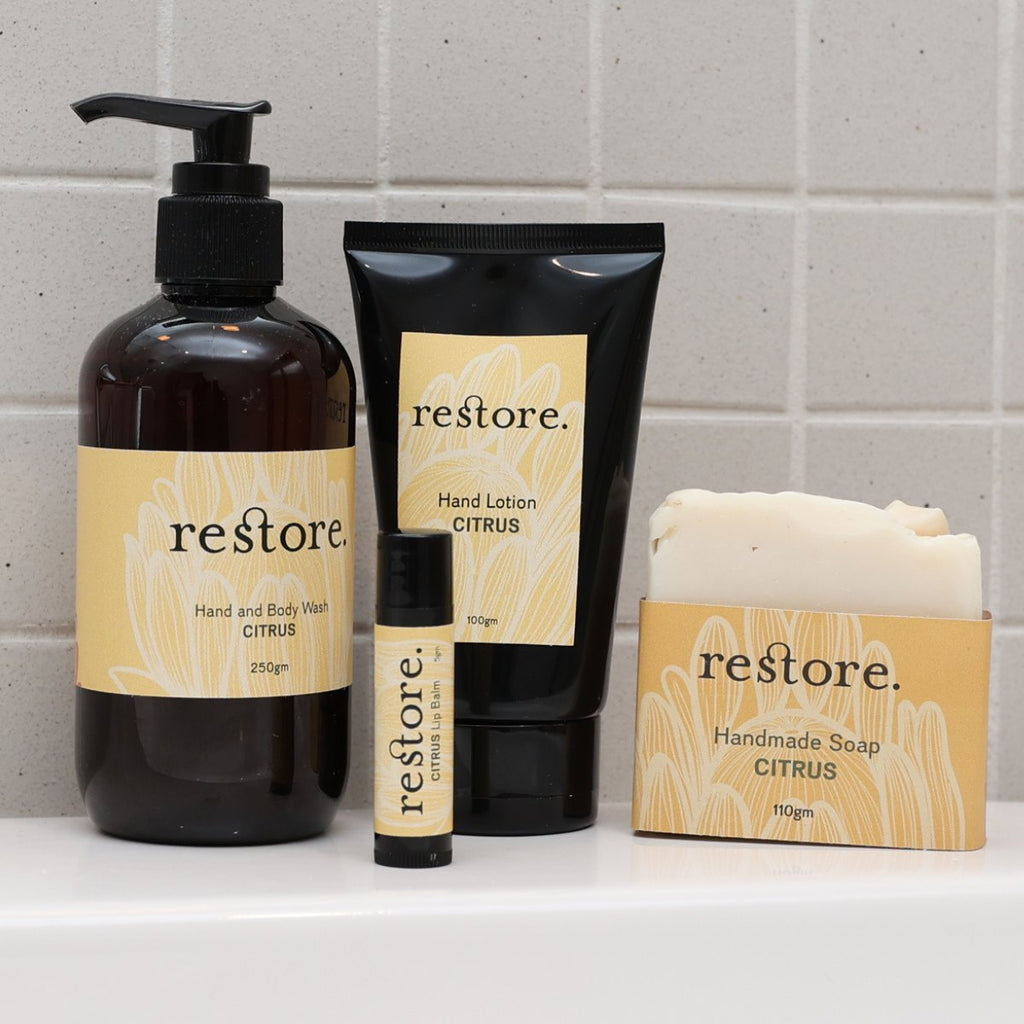 A set of four 'restore.' citrus-scented bath and body products arranged on a white ledge against a light gray tiled wall: a dark amber pump bottle, a black hand lotion tube, a small black lip balm tube, and a bar of handmade soap in a matching box.