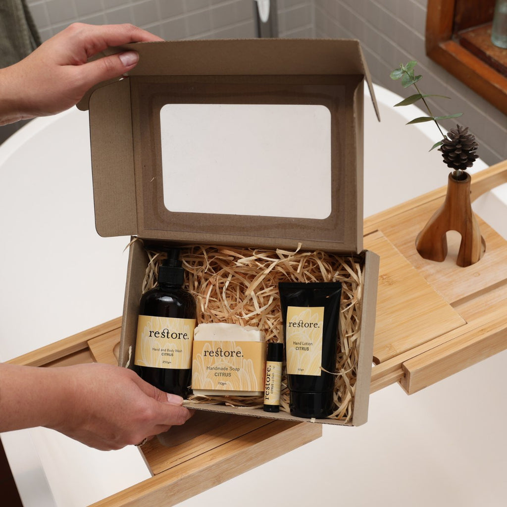 A person holds open a brown cardboard gift box with a clear window, containing bath products on a wooden caddy beside a bathtub. Visible products include a black bottle labeled restore, a cream tube labeled restore, a small tube, and a wrapped item, all arranged in shredded paper filler.