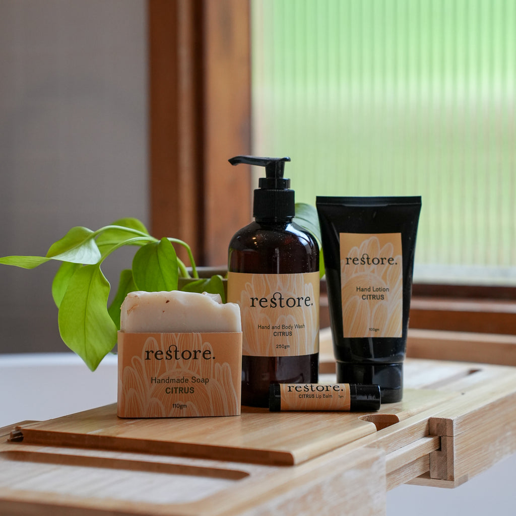 A set of restore. personal care products including handcrafted soap, a pump bottle, a tube, and a small tube, arranged on a wooden tray with a green plant in front of a window.
