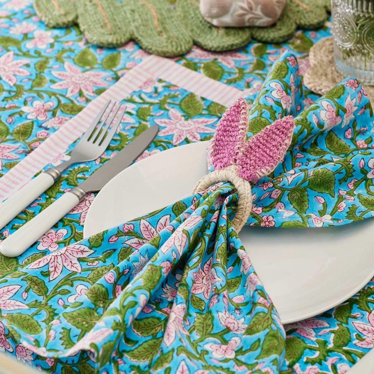 Table setting with floral tablecloth, pink butterfly napkin holder, and cutlery.