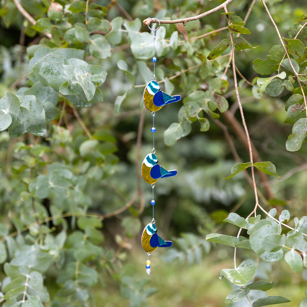 Three hanging bird-shaped glass wind chimes in blue, yellow, and white, suspended from a tree branch with green leaves in the background.