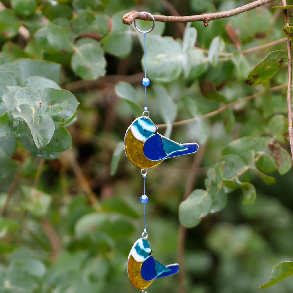 A hanging stained glass bird wind spinner with two birds, blue beads, and a metal ring, photographed outdoors against a blurred background of green leaves.