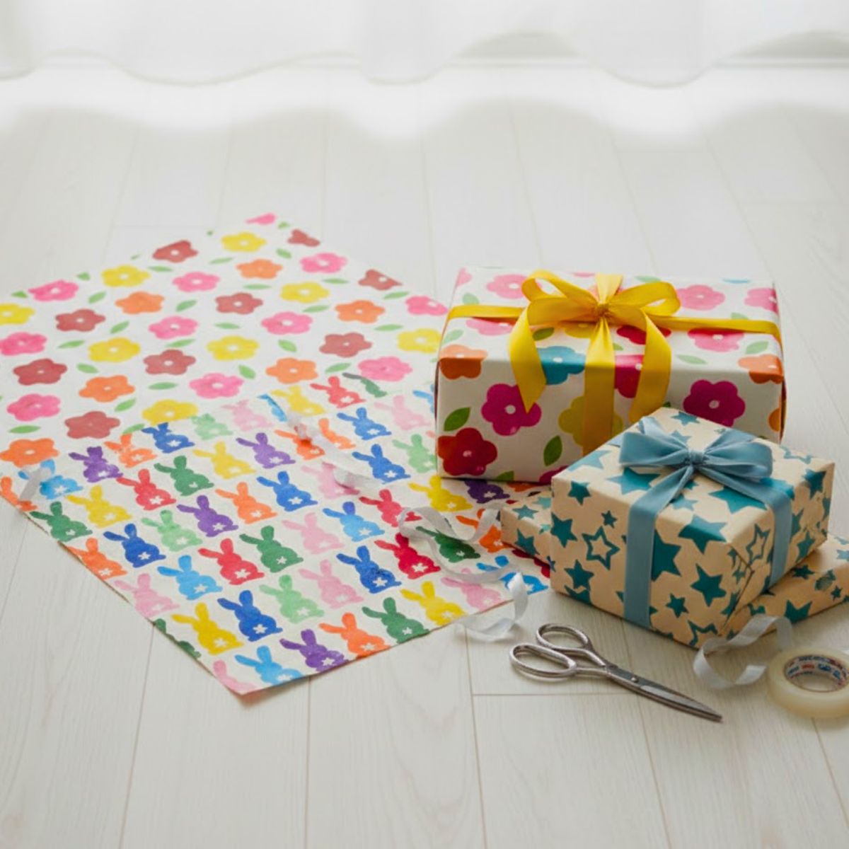 Colorful gift wrap with floral and star patterns, ribbons, scissors, and tape on a white surface.