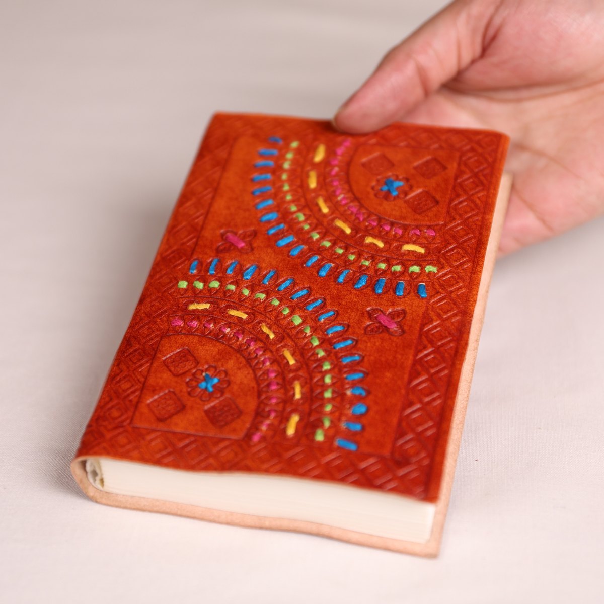A hand holding a small leather journal with an orange-brown cover decorated with colorful embroidered geometric patterns, on a light beige background.