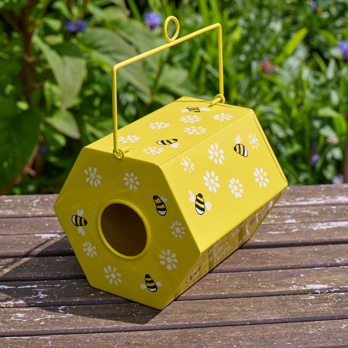 Yellow bee-themed birdhouse with floral and bee designs on a wooden surface with a green garden background.