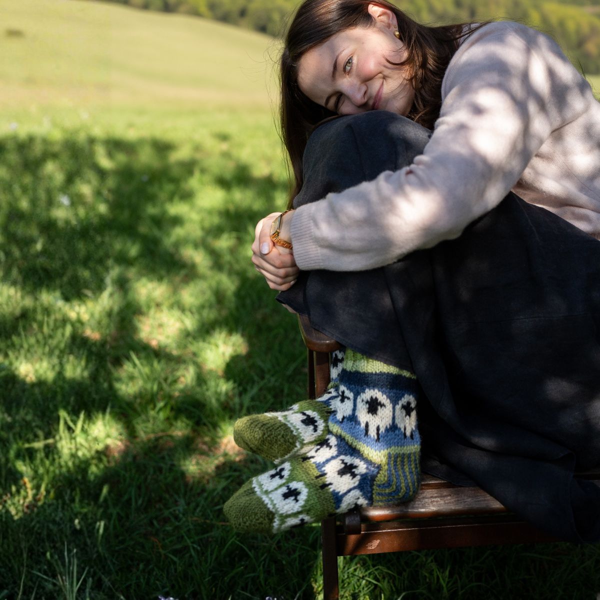 A women sitting on a wooden chair in a grassy field with knitted socks.