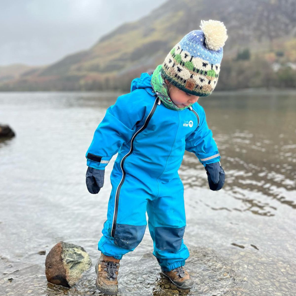 Child in a blue waterproof suit and colorful hat standing by a body of water with mountains in the background.