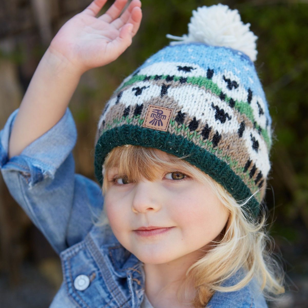 Child wearing a colorful knit hat with a brand logo, waving outdoors.