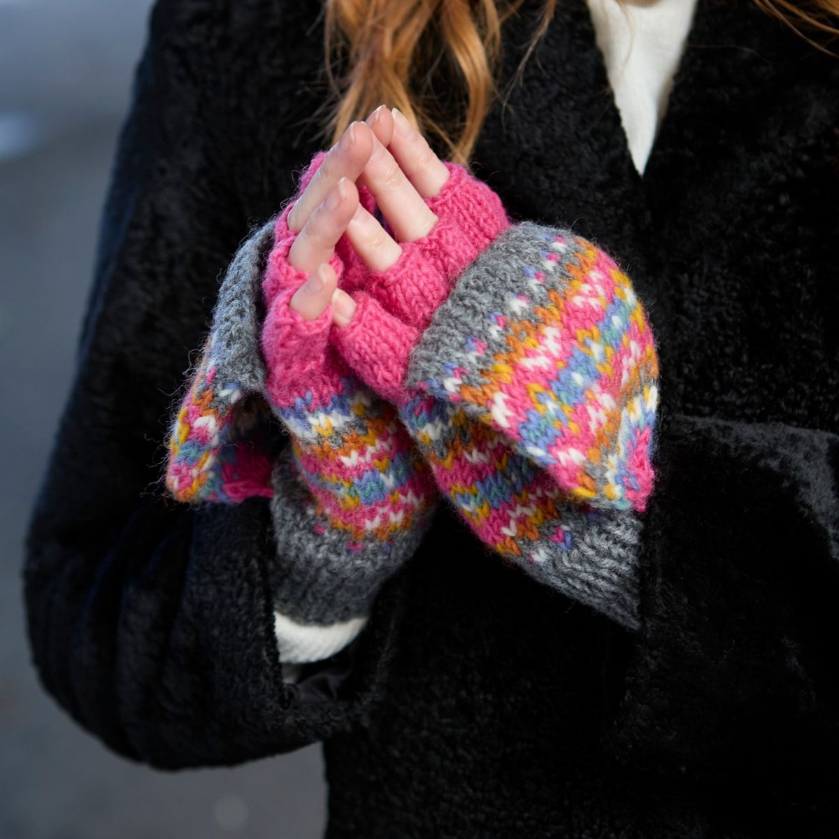 Colorful knitted fingerless gloves held by a person wearing a black coat.