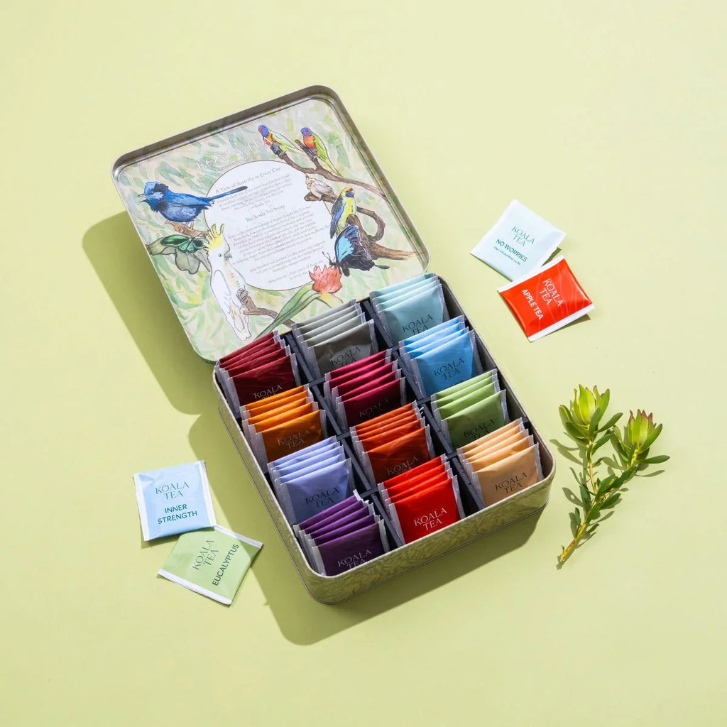 Open tin of assorted tea bags with colorful packaging on a light yellow background