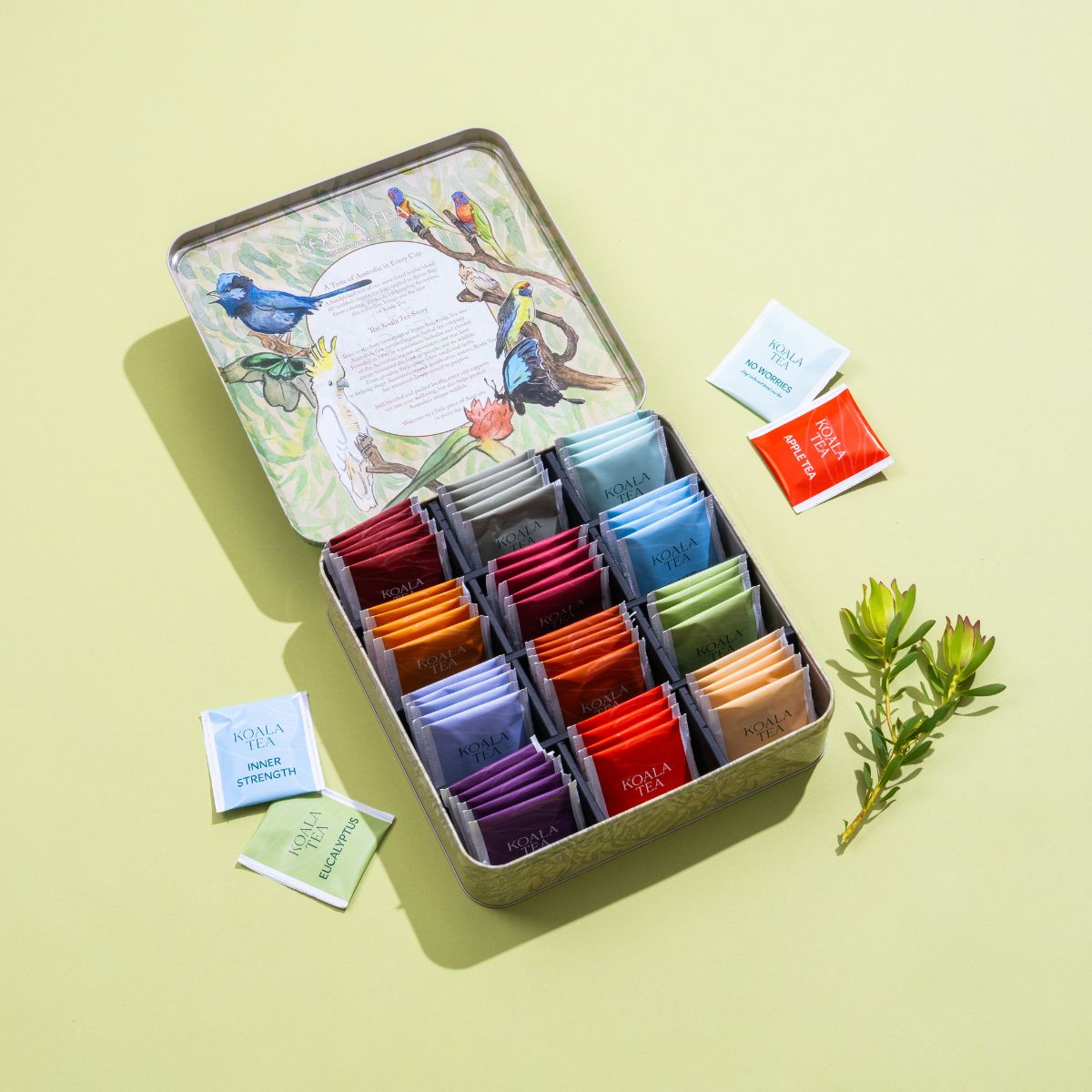 Open tin of assorted tea bags with colorful packaging on a light yellow background