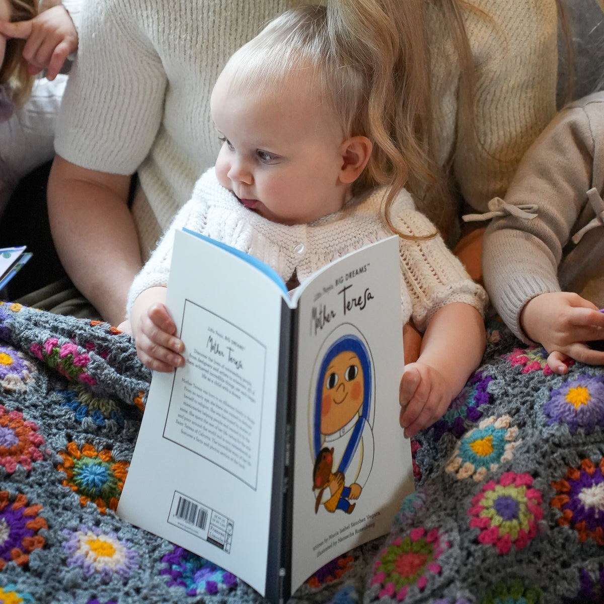 A toddler sits on a crocheted blanket reading a children's book titled 'Mile High' with a smiling character illustration on the cover, surrounded by adults in a cozy indoor setting.