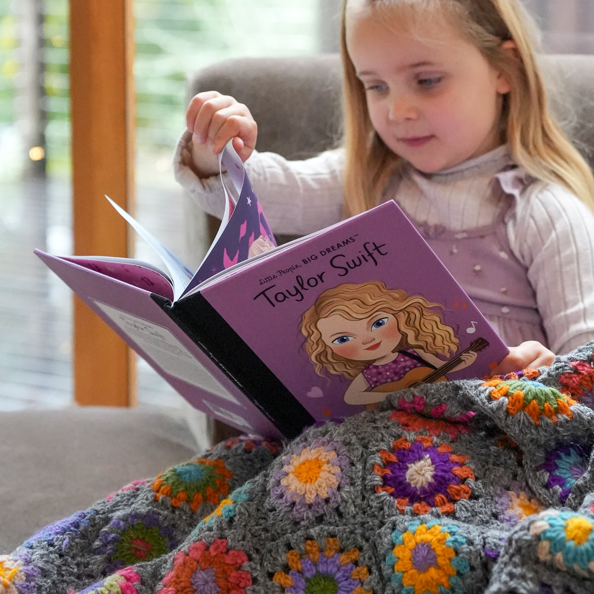 A young girl sits in a chair reading a purple children's book titled 'Taylor Swift' under a gray crocheted blanket with colorful floral patterns.