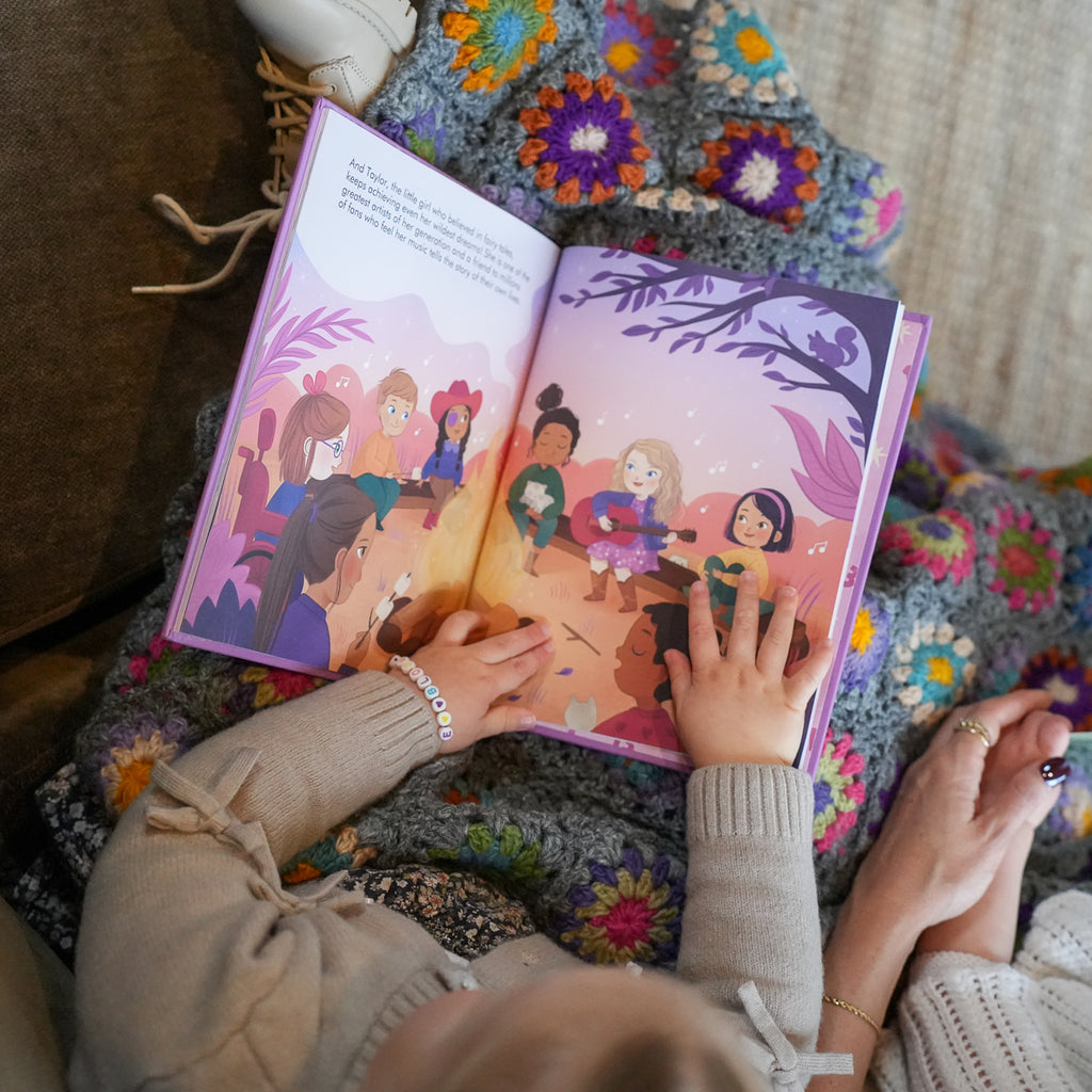 A child's hands holding an open children's picture book with colorful illustrations of children sitting on a cozy, multicolored crocheted blanket.