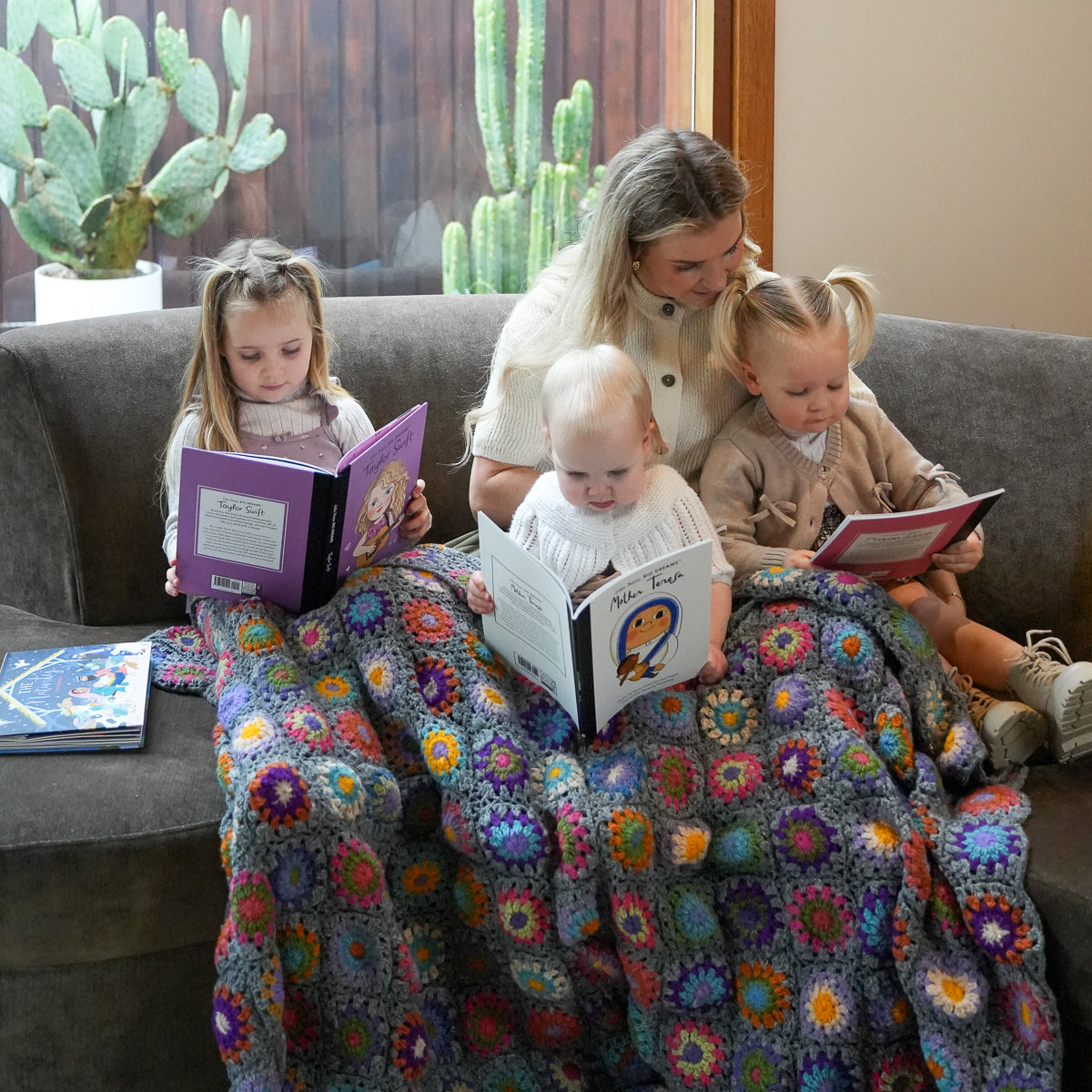 A woman and three children sit on a gray couch reading books, wrapped together in a large crocheted blanket with a multicolor granny-square pattern. A potted cactus is visible in the background near a window.