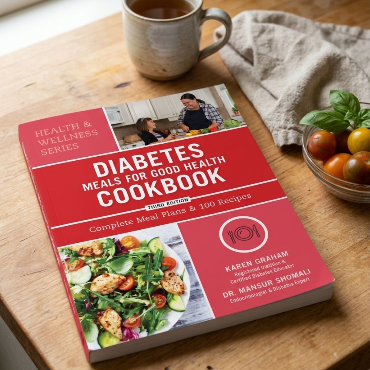 Cookbook titled 'Diabetes Meals for Good Health Cookbook' on a wooden table with a cup of tea and tomatoes.