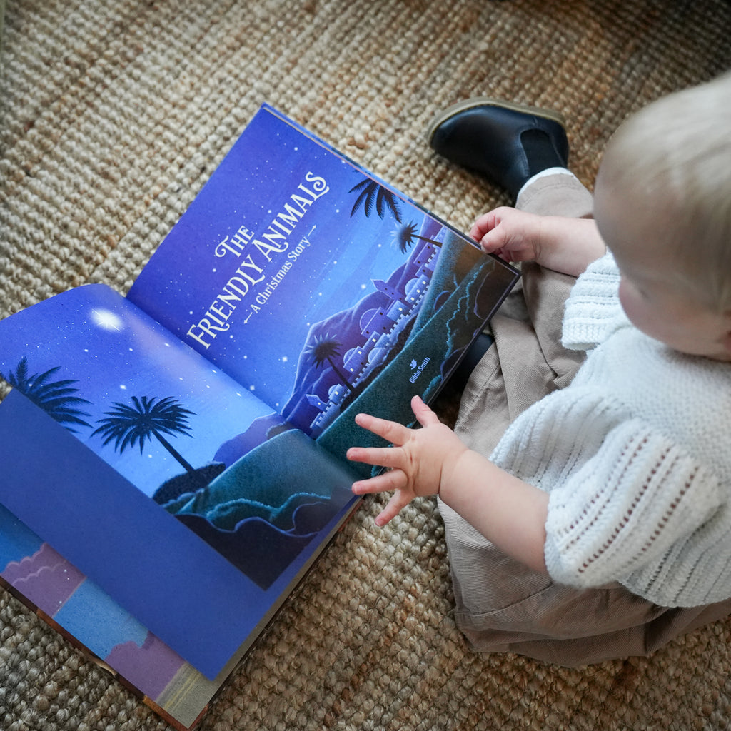 A baby sitting on a woven rug reading a colorful children's picture book titled 'The Friendly Animals: 20 Gentle Stories'. The book features a vibrant blue night sky and landscape illustration with palm trees.