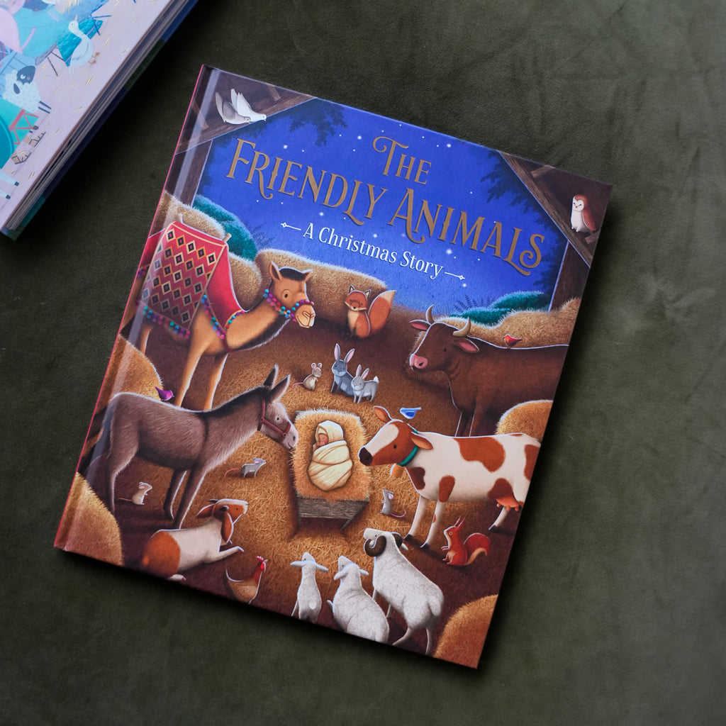 A hardcover children's book titled "The Friendly Animals: A Christmas Story" lies on a dark textured surface. The cover features a colorful illustration of various animals gathered around a baby in a manger, with a deep blue night sky at the top.