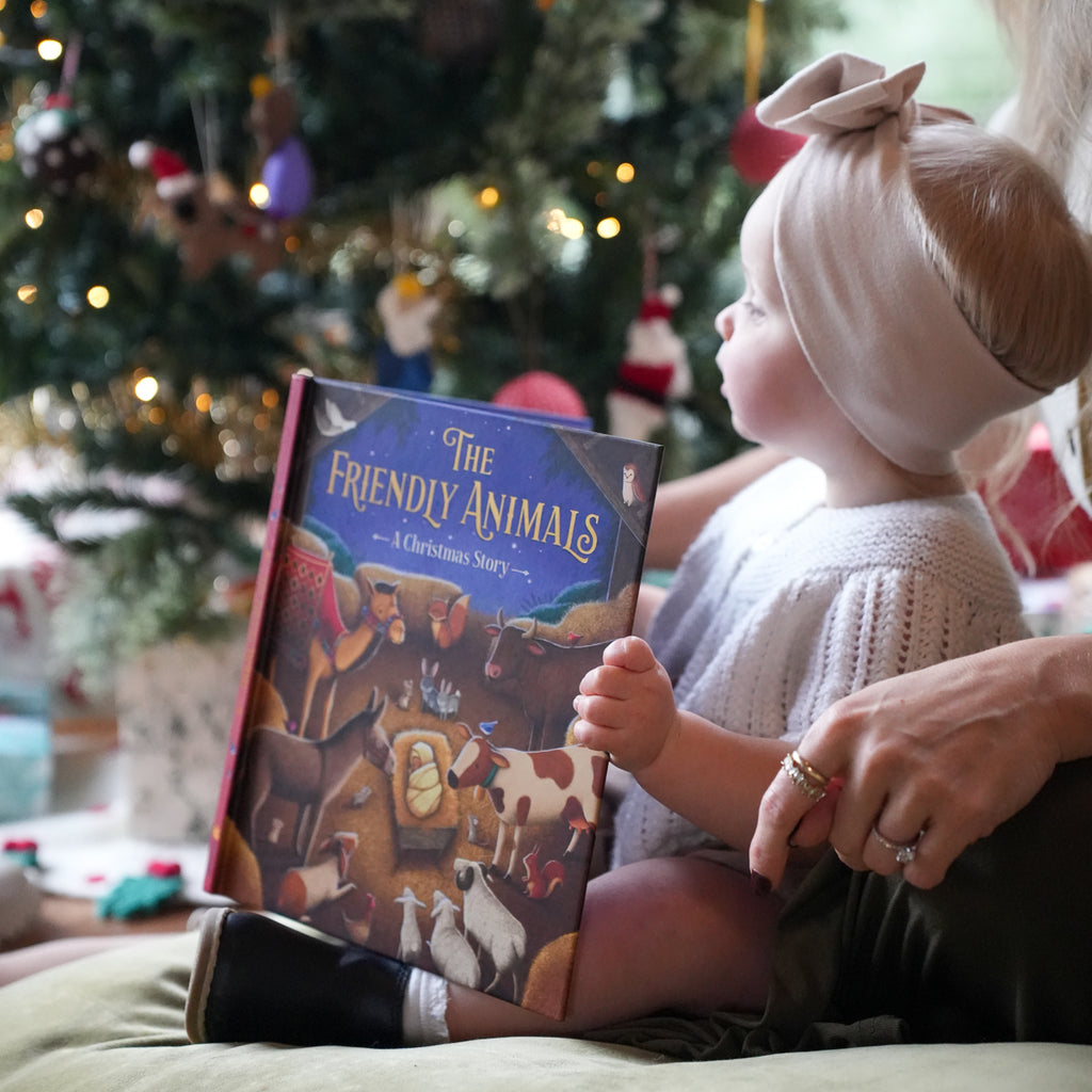 A young child in a white knit sweater and beige headband sits beside an adult, holding a hardcover book titled "The Friendly Animals: A Christmas Story" in front of a softly blurred Christmas tree with warm lights.