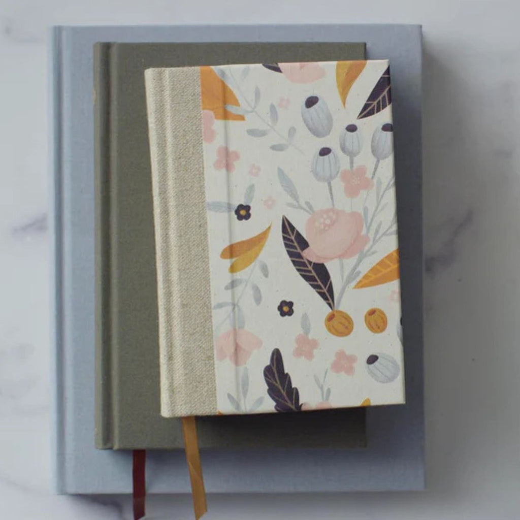 A floral patterned hardcover notebook with a beige fabric spine and ribbon bookmark, resting on top of a muted sage green book, which is placed on a light blue book against a white marble background.
