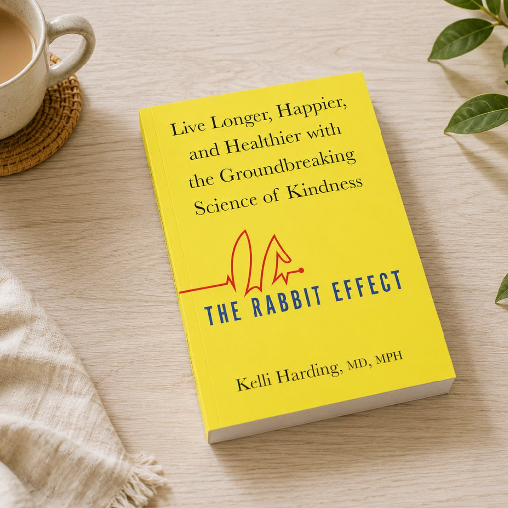 A yellow book titled "The Rabbit Effect" by Kelli Harding, MD, MPH, placed on a light wood surface with a cup of coffee, a woven coaster, and green leaves nearby.