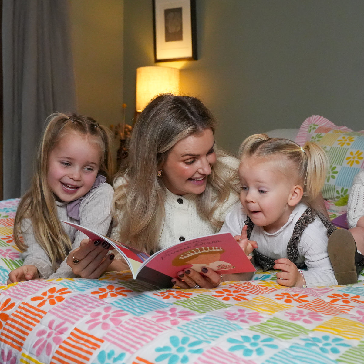 A woman and two young girls are lying on a bed reading a colorful book. The bed is covered with a bright, patterned quilt in pink, blue, yellow, and orange.