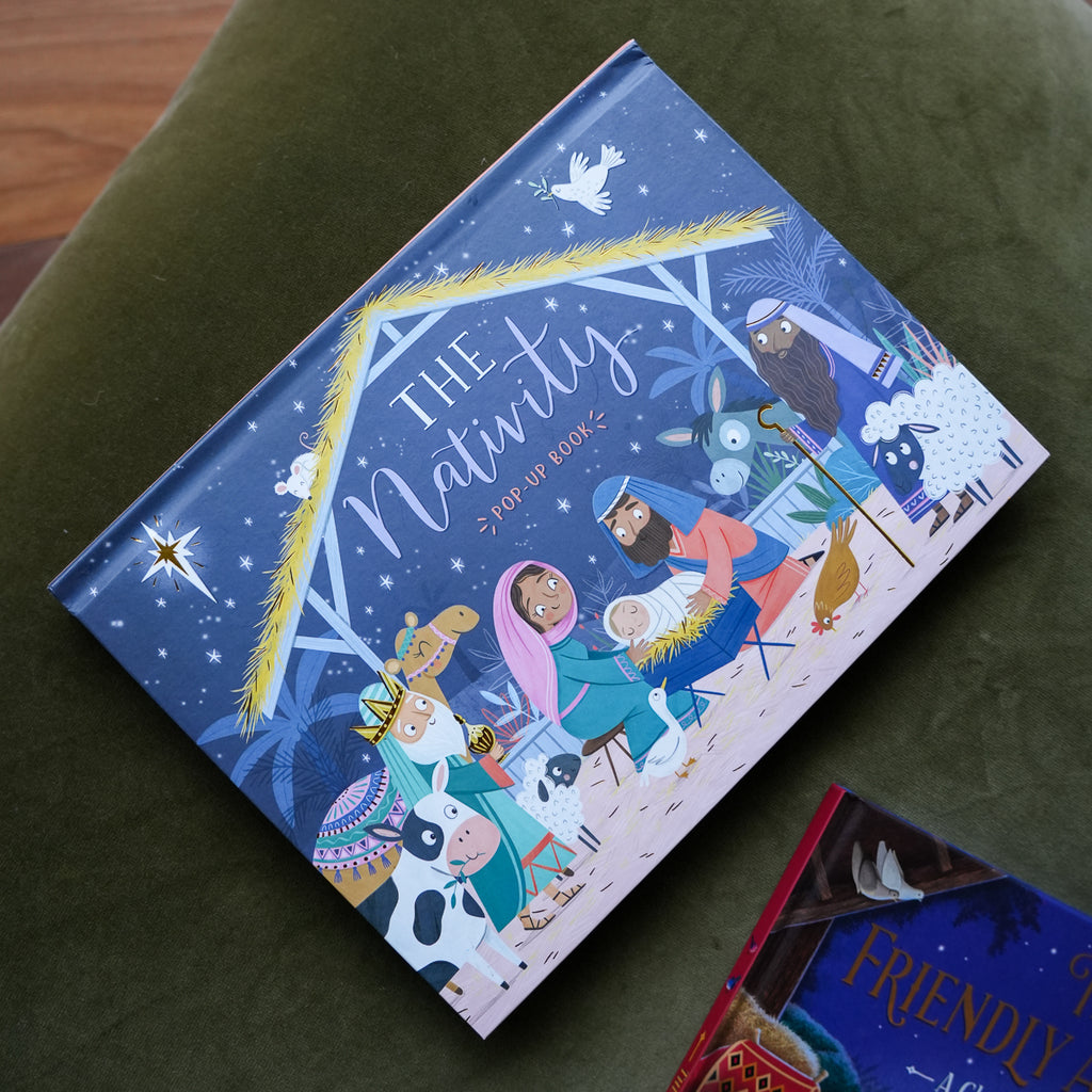 A hardcover book titled 'The Nativity' featuring a colorful, whimsical nativity scene illustration on the cover, resting on an olive green velvet cushion.