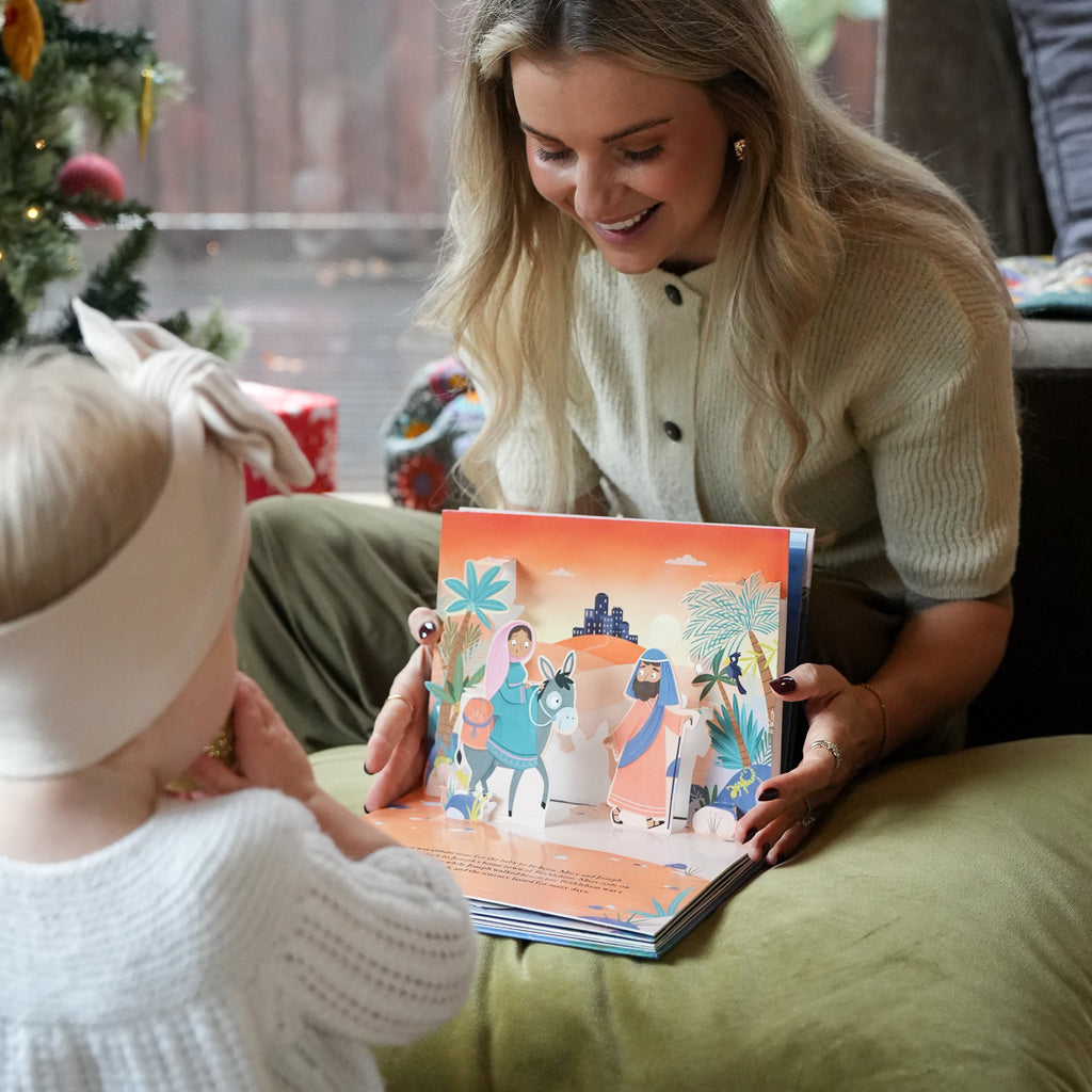 A woman and a baby are sitting together indoors, looking at a colorful 3D pop-up children's book. The book features a desert scene with paper-cut characters.