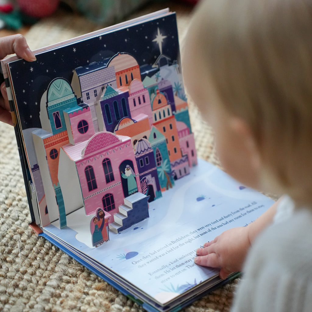 A baby is reading a colorful 3D pop-up children's book about the story of Mary and Joseph, held open by an adult's hand on a textured beige rug.